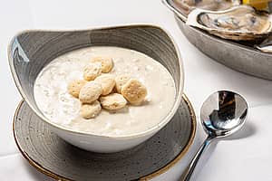 Bowl of New England clam chowder with oyster crackers on top in bowl with spoon to right.