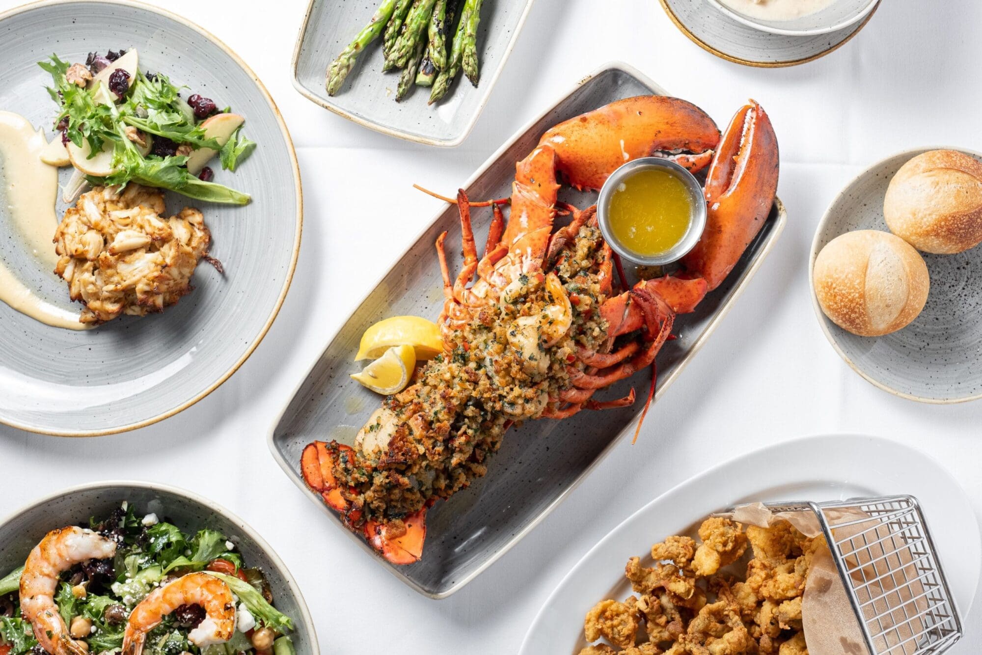 A variety of seafood dishes served on a table. A whole lobster with a small container of melted butter, asparagus, crab cakes with a small salad, garlic shrimp, bread rolls, and fried seafood in a small basket are arranged on white plates.