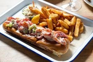 Half pound Maine lobster roll with lemon mayo, french fries and coleslaw on metal tray.