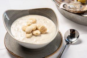 Bowl of New England clam chowder with oyster crackers on top in bowl with spoon to right.