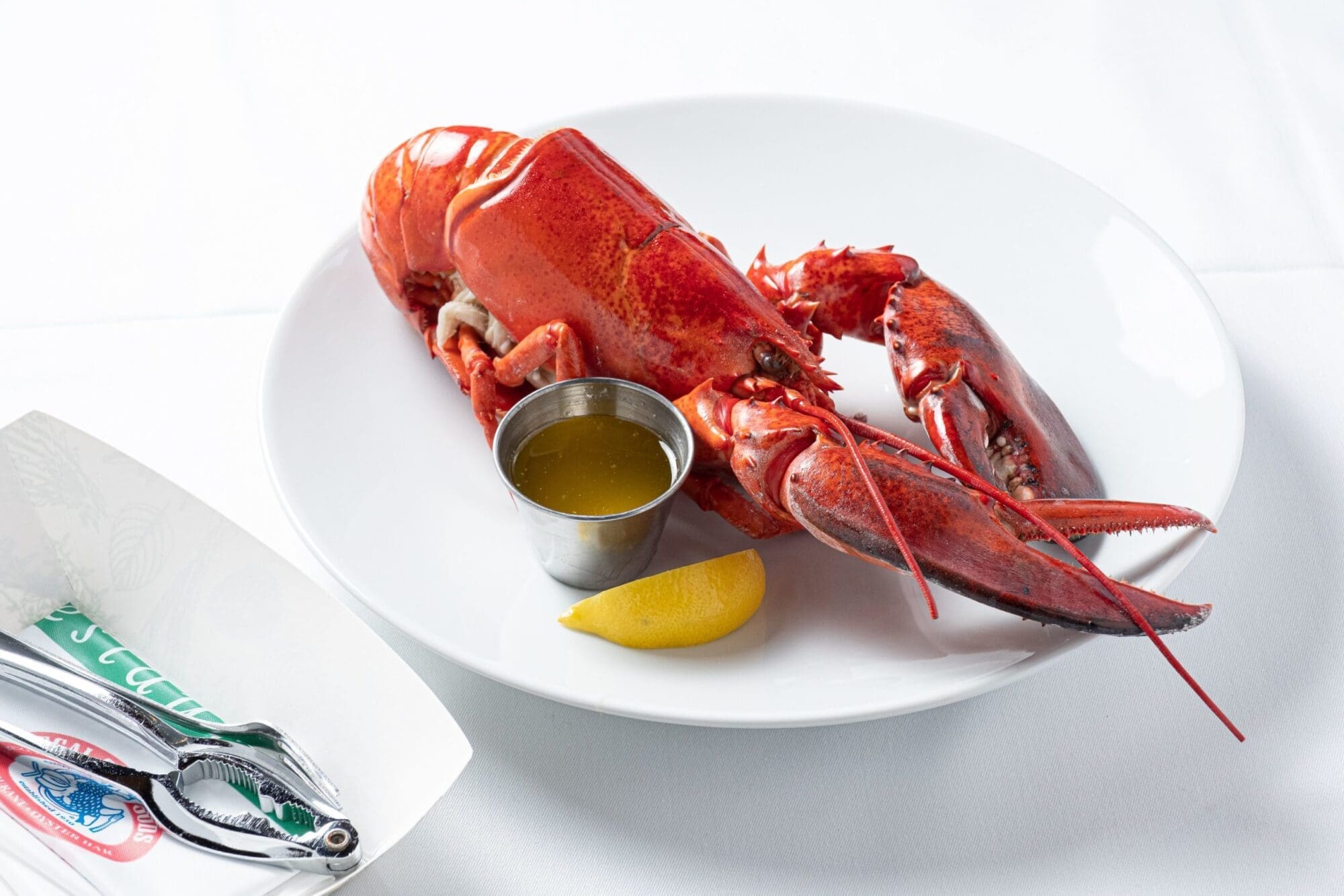 A whole cooked lobster is served on a white plate with a small stainless steel cup of melted butter and a lemon wedge. A lobster cracker tool and bib are placed on the left side of the image.