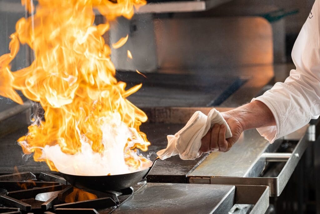 A chef's hand holding a frying pan with a large flame rising from it on a stovetop. The chef is holding a towel in the other hand, and the kitchen background is blurred.