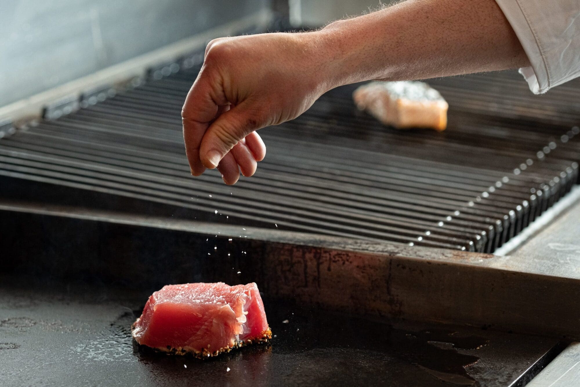 Chef sprinkling sale on raw tuna that was just set on the grill to cook.