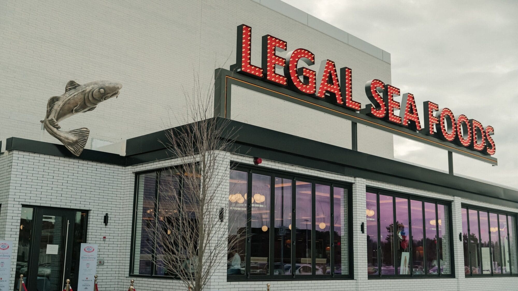 Exterior of a Legal Sea Foods restaurant with a large red and white sign on a brick building. A fish decoration is mounted on the wall near the sign. There are large windows and a tree in front of the building under a cloudy sky.