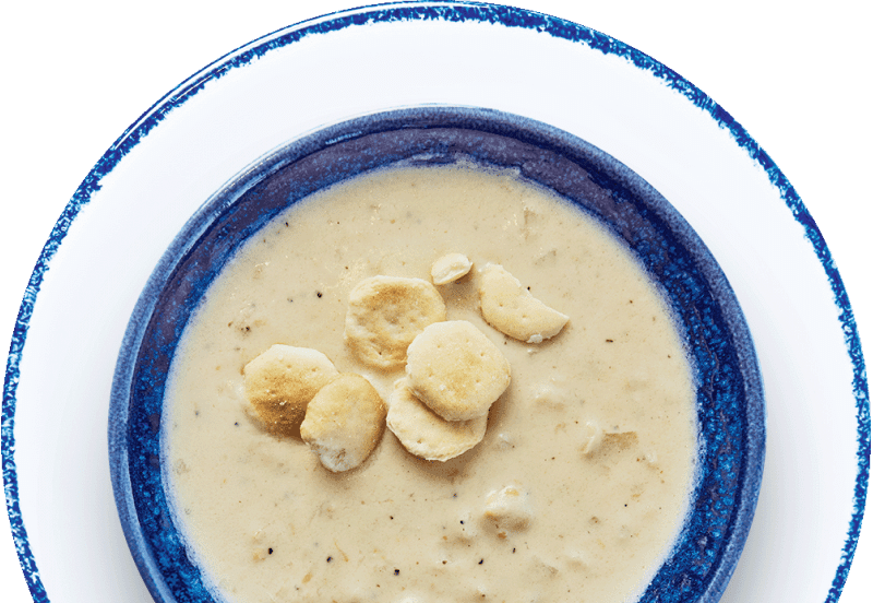 A blue ceramic bowl filled with creamy soup, topped with several round, crispy crackers. The bowl is placed on a white plate with a blue rim. The soup has a thick texture and is light beige in color.