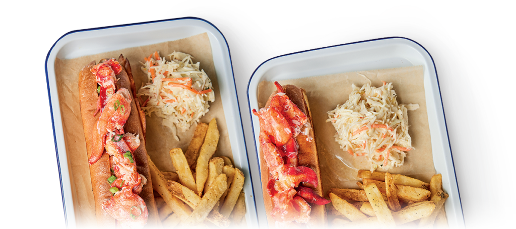 Two rectangular trays each contain a lobster roll in a toasted bun, a serving of seasoned fries, and a portion of coleslaw. The rolls are filled with lobster meat mixed with a light dressing. The trays are lined with parchment paper.