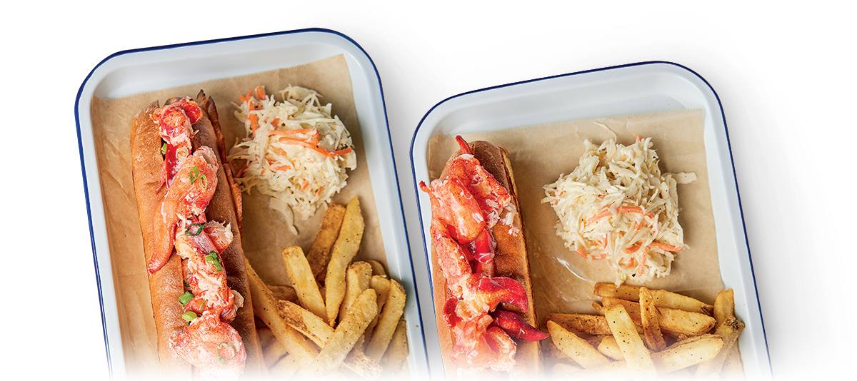 Two rectangular trays each contain a lobster roll in a toasted bun, a serving of seasoned fries, and a portion of coleslaw. The rolls are filled with lobster meat mixed with a light dressing. The trays are lined with parchment paper.