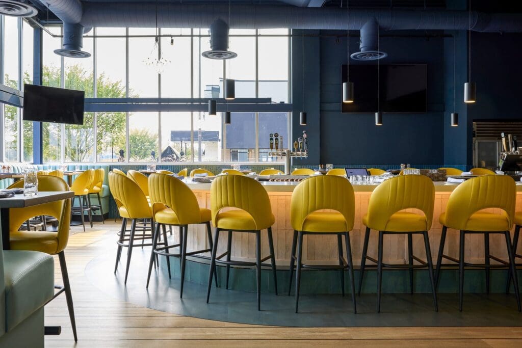 A modern bar with a circular counter surrounded by bright yellow chairs, large windows letting in natural light, and multiple flat-screen TVs mounted on dark blue walls.