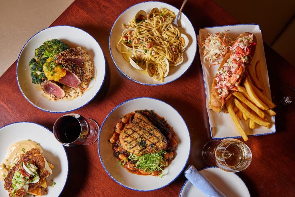 A wooden table with five dishes: a bowl of pasta with clams, seared tuna with greens, a lobster roll with fries, grilled fish on vegetables, and a creamy chicken dish. Two wine glasses, one with red wine and the other with white wine, are beside the plates.
