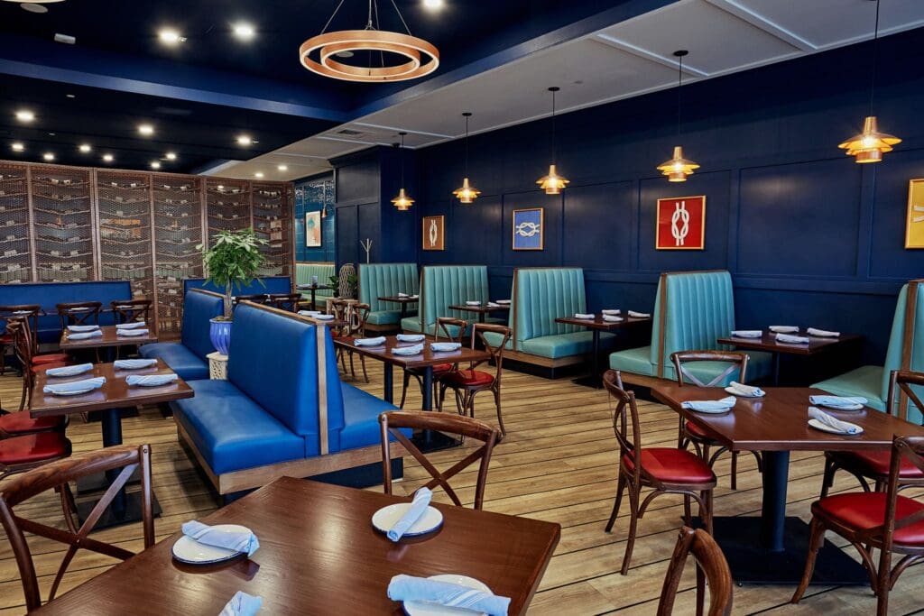 Cozy restaurant interior with wood flooring and dark blue walls. Tables are set with white napkins on dark wood surfaces. Green and blue booth seating along the walls, accented by warm pendant lights and modern art pieces.