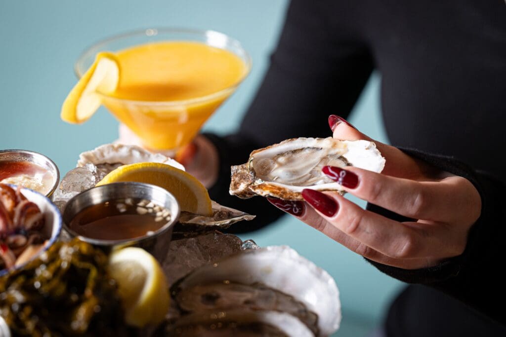 A person with long red nails holds an open oyster. There is a seafood platter with oysters, lemon wedges, and sauces on ice. A yellow cocktail with a lemon slice is in the background.