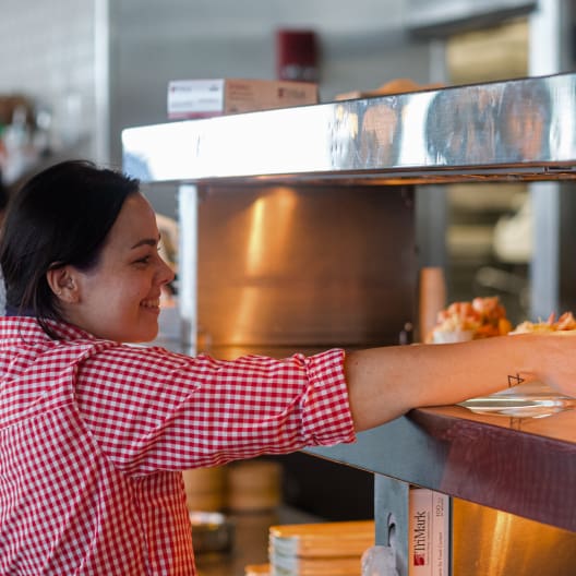 A smiling person wearing a red and white checkered shirt is placing a plate of food on a counter in a restaurant kitchen. The background shows kitchen equipment and ingredients.