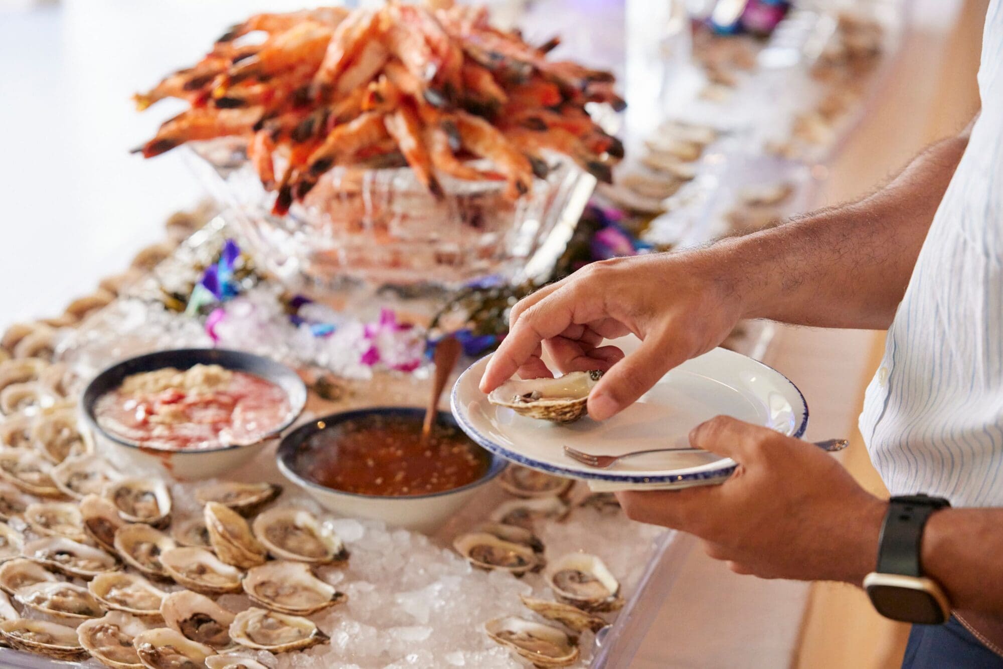A person is holding a plate and preparing to eat an oyster from a seafood buffet. The table displays a variety of shellfish, including a large dish of shrimp and bowls of sauces, all set on crushed ice.