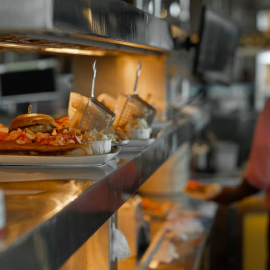 Plates of food, including a burger with fries and dishes in small wire baskets, are placed on a stainless steel counter under a warming light in a busy restaurant kitchen, with a blurred figure in the background.