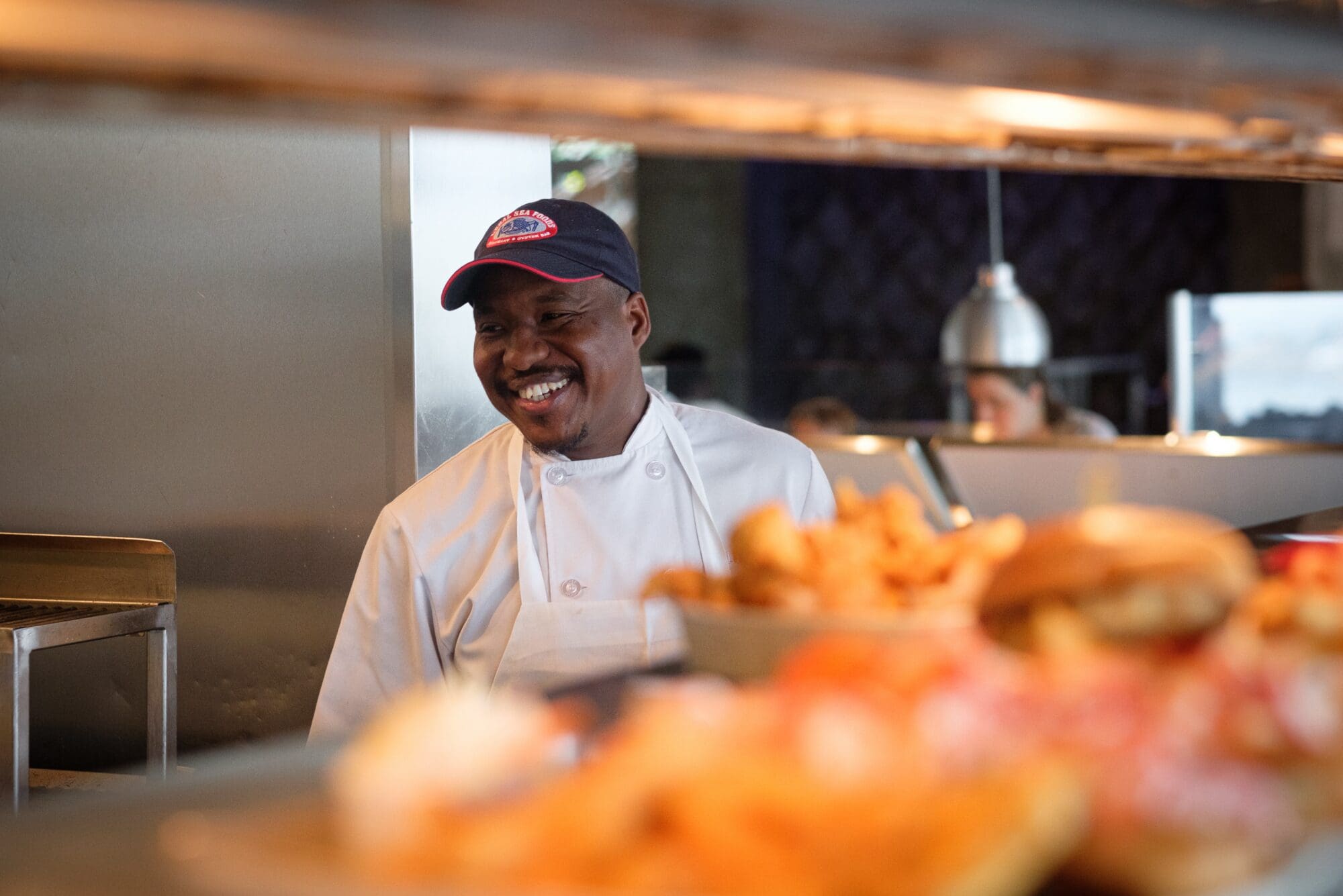 A smiling chef in a white uniform and a baseball cap stands behind a kitchen counter with various plated dishes in the foreground.