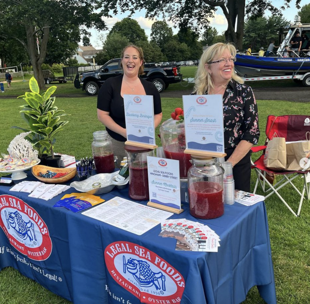 Two smiling women stand behind a blue Legal Sea Foods table at an outdoor event. The table displays drink dispensers, brochures, business cards, and a plant. A black truck and a blue and yellow boat are visible in the background on the green grass.