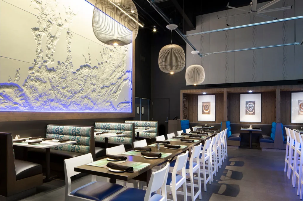 Modern restaurant interior with a large map-like wall art illuminated in blue lights. The dining area has booth seating with teal and patterned upholstery, white chairs, and a long table decorated with black plates. Contemporary light fixtures hang from the ceiling.