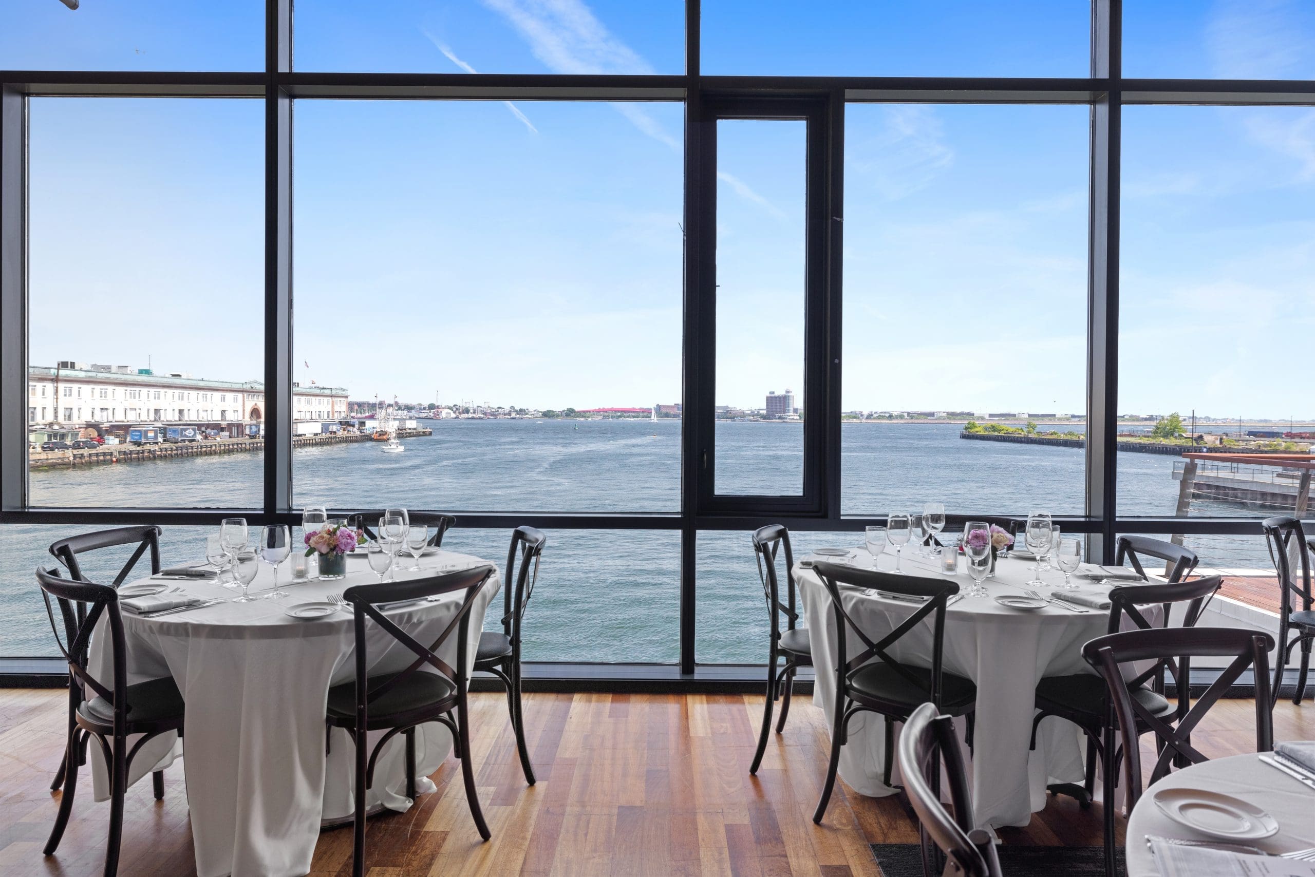 Restaurant dining room with tables set for a meal, featuring white tablecloths and tableware. Large windows offer a scenic view of a waterfront with calm water and distant buildings under a clear blue sky.