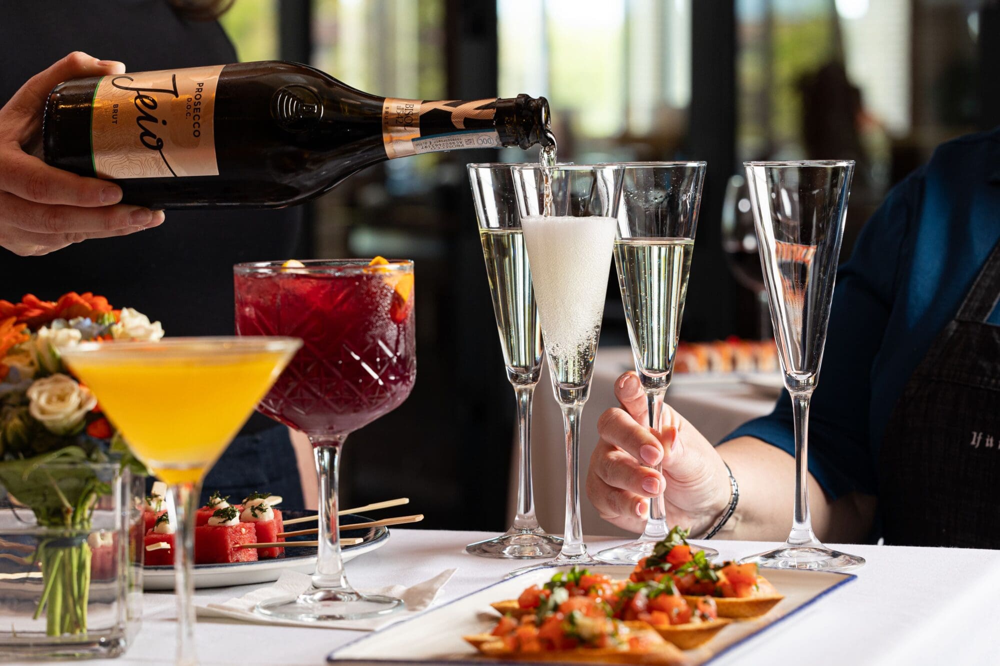 A hand pours Prosecco into a champagne flute at a table with various cocktails, including a yellow drink and a red sangria. Appetizers like bruschetta are on plates. The scene suggests a celebratory or elegant dining setting.