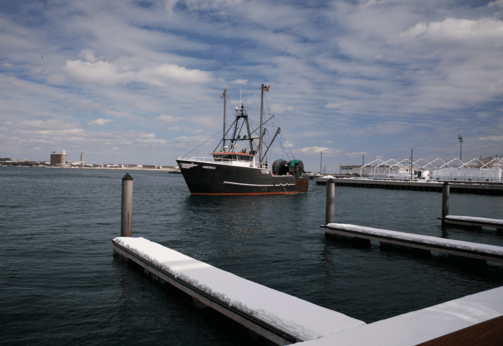 A fishing boat is docked in a harbor. The boat has black and red detailing and is equipped with fishing nets and equipment. The harbor features snowy docks and calm water under a partly cloudy sky. White buildings and distant structures can be seen in the background.