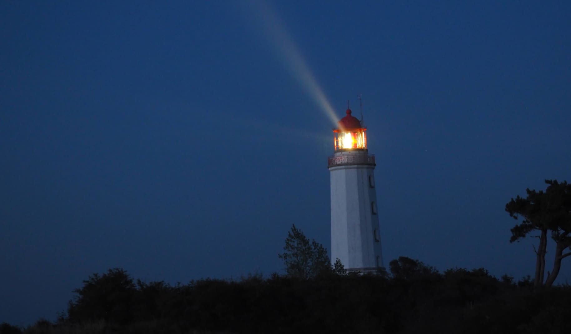 A lighthouse stands tall and illuminated against the night sky. A bright beam of light shines from the top, cutting through the dark blue of the evening. The surrounding area is covered in silhouetted trees and bushes.