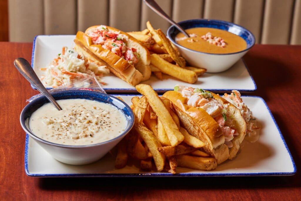 Two plates with lobster rolls, French fries, and coleslaw; each is paired with a bowl of creamy white or orange soup. Enjoy this Express Lunch Combo at Legal Sea Foods, all served on a wooden table.
