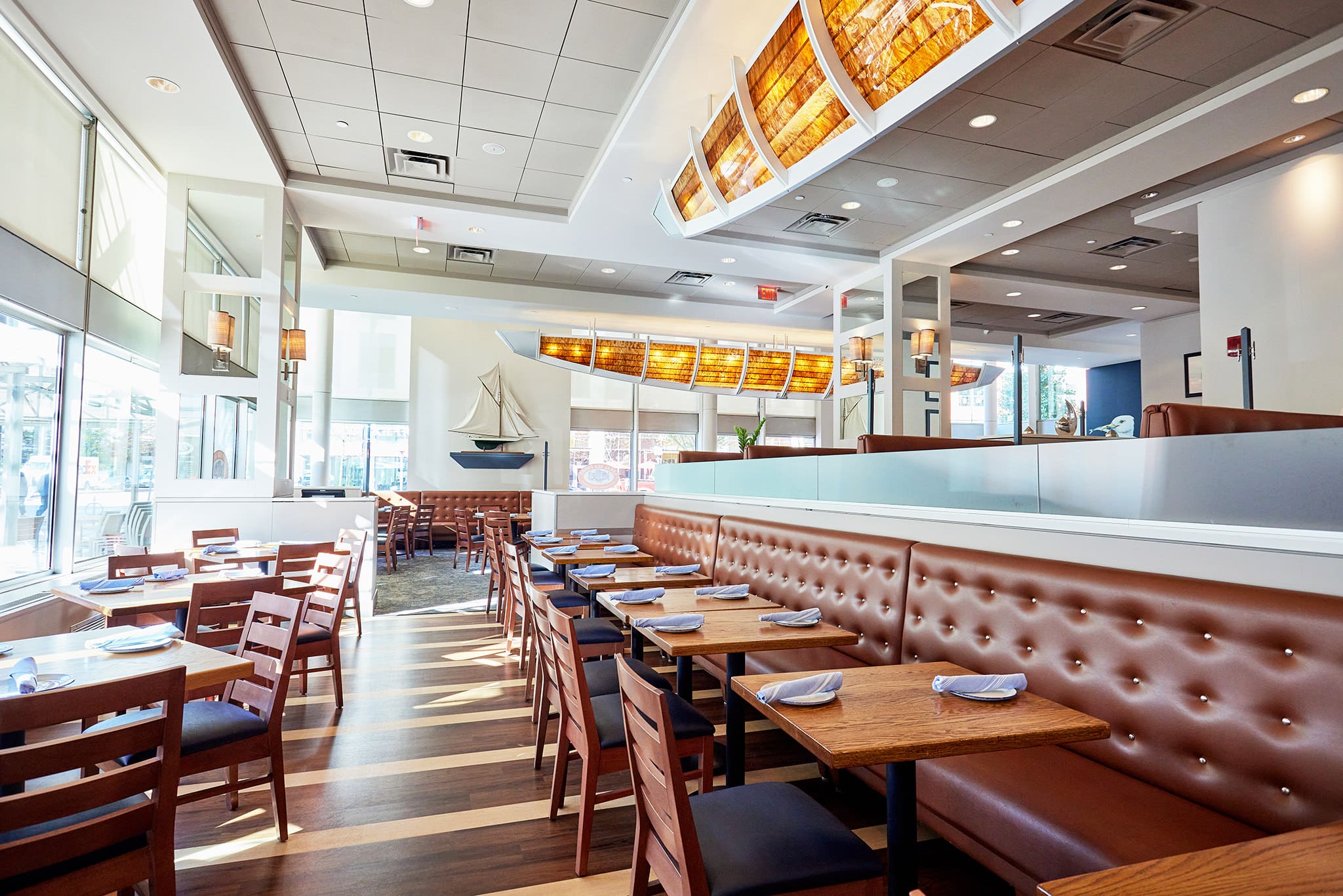 A modern restaurant interior with wooden tables and chairs, brown upholstered booth seating, and large windows letting in plenty of natural light. Decorative elements include a boat sculpture on the far wall and rectangular ceiling lights with warm tones.
