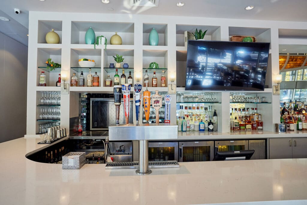 A modern bar with a sleek white countertop features a wall of shelves holding various bottles of liquor, glasses, and decorative items. A row of beer taps is in front of the shelves, and a large flat-screen TV is mounted on the right side of the shelving unit.