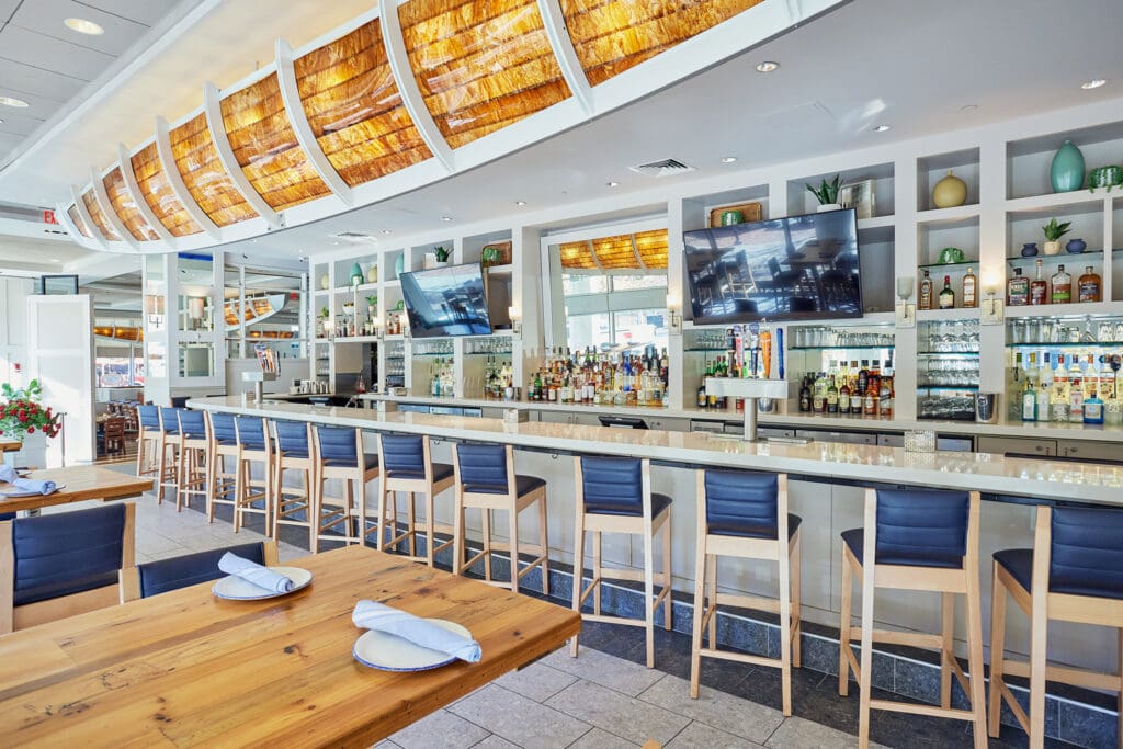 Modern bar interior with a long countertop, high bar stools, two large flat-screen TVs, and a well-stocked selection of beverages behind the bar. The bar is well-lit with warm, decorative ceiling lights and features a contemporary design with light wood and blue accents.