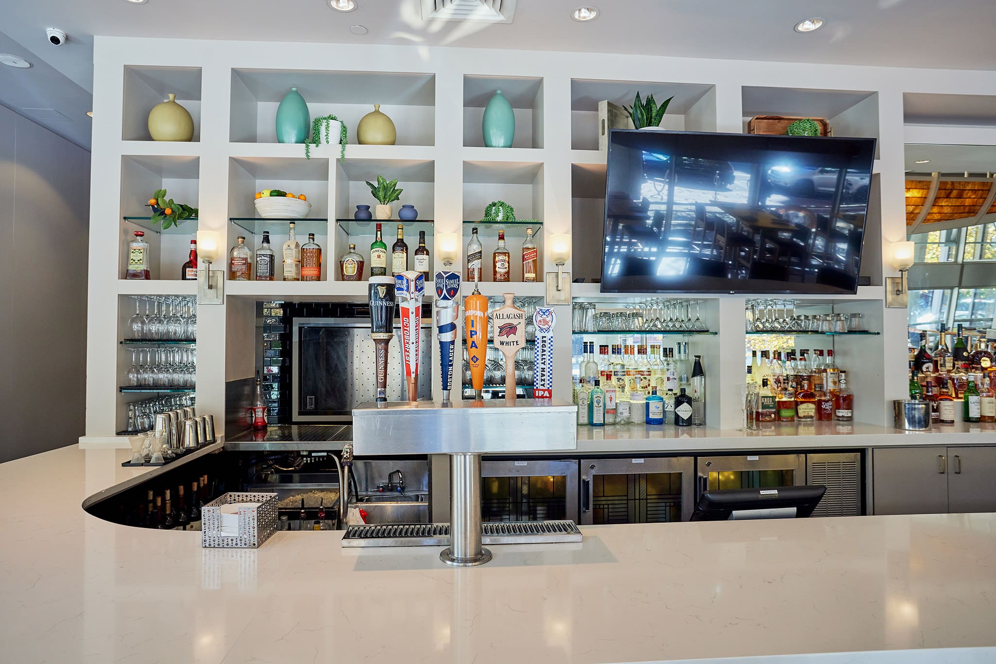 A modern bar with a sleek white countertop features a wall of shelves holding various bottles of liquor, glasses, and decorative items. A row of beer taps is in front of the shelves, and a large flat-screen TV is mounted on the right side of the shelving unit.