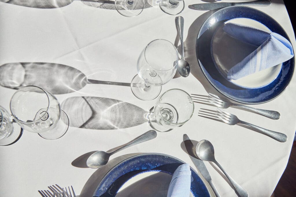 A table set for two with white tablecloth, blue-rimmed plates, folded napkins, and silver cutlery. Four wine glasses are placed near the plates, casting shadows on the table. The setup gives a formal dining impression.