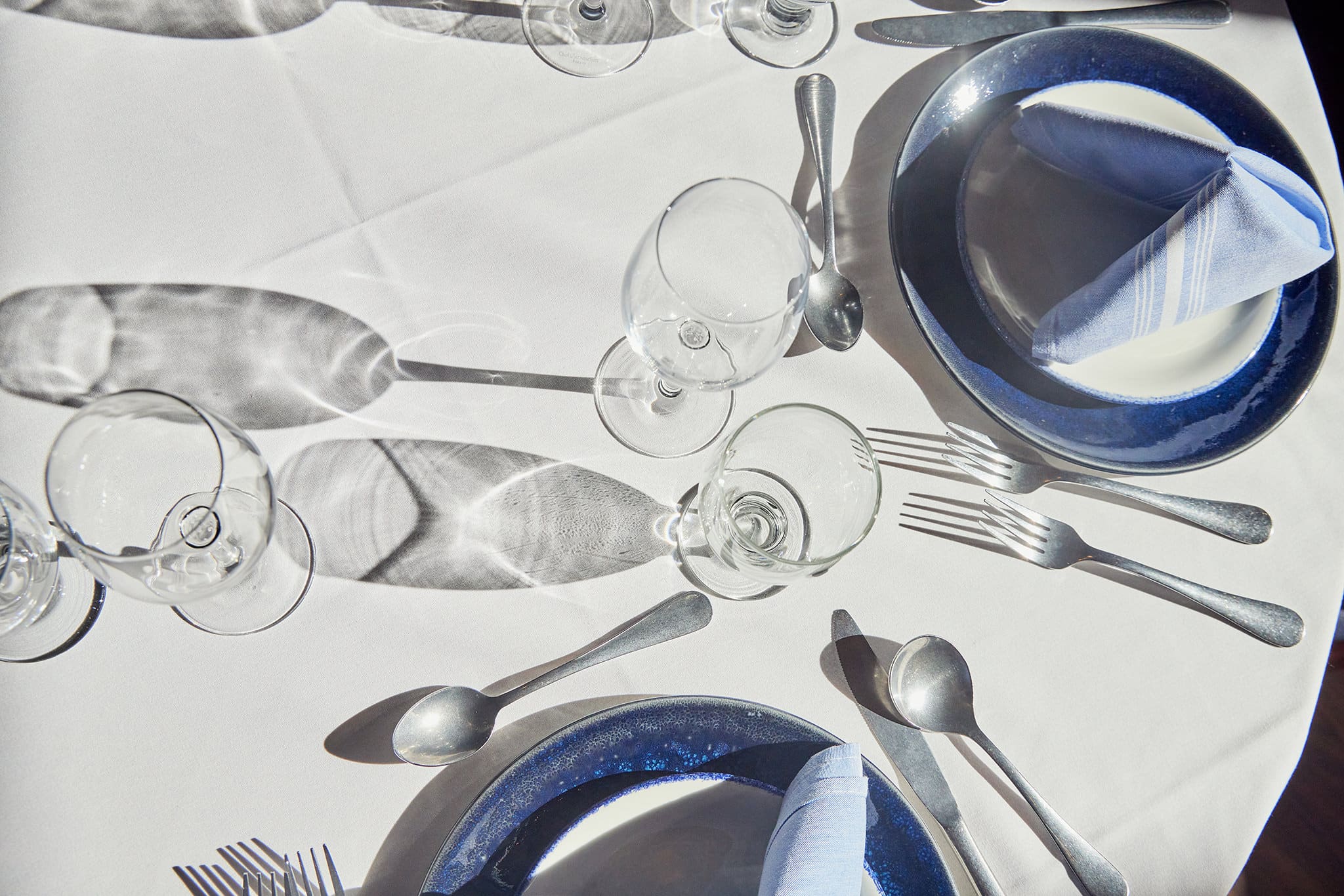 A table set for two with white tablecloth, blue-rimmed plates, folded napkins, and silver cutlery. Four wine glasses are placed near the plates, casting shadows on the table. The setup gives a formal dining impression.