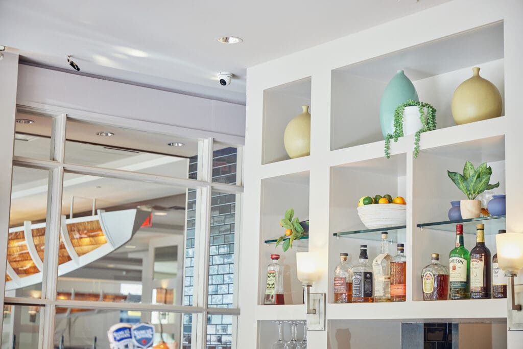 A modern bar interior with white shelves displaying various bottles of alcohol, along with decorative vases and plants. The background features glass walls with a view of additional seating and a ceiling with overhead lights.