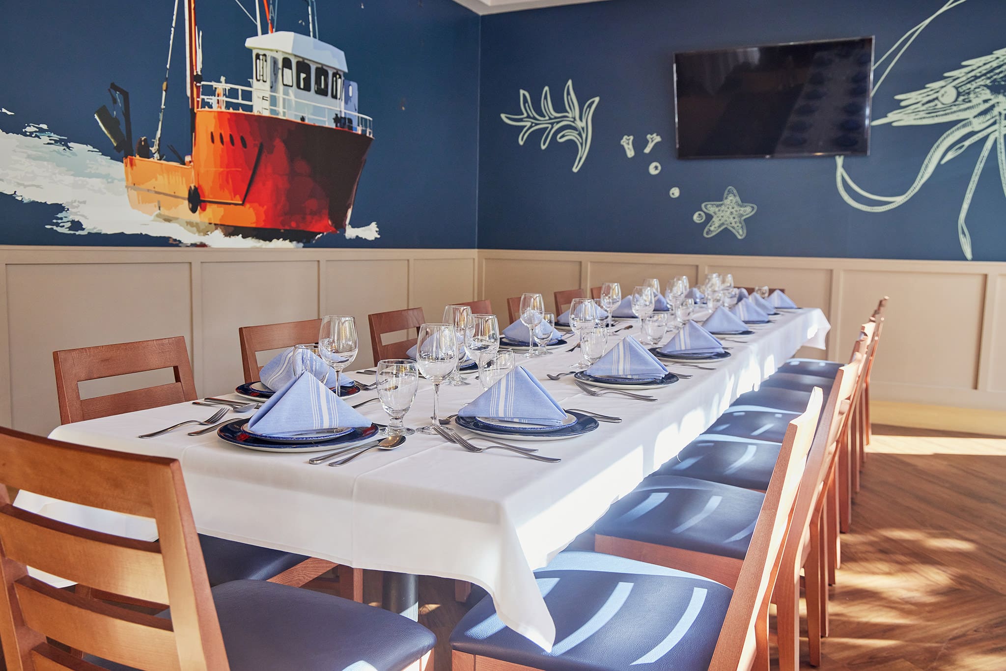 A long dining table set for a meal with neatly folded blue napkins, glasses, and cutlery in a room featuring a large mural of a fishing boat and sea creatures on the wall. There is a flat-screen TV mounted on the wall, and wooden chairs surround the table.