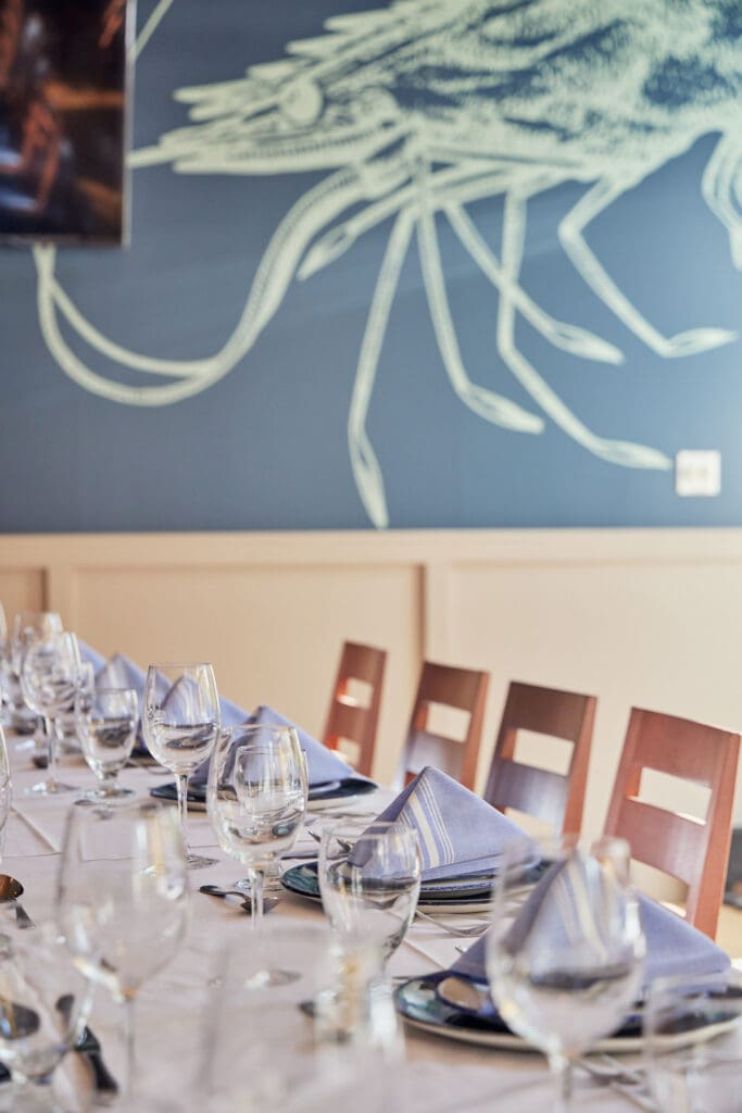 A restaurant table set for a meal with neatly arranged wine glasses, plates, and folded napkins. Wooden chairs line the table, and a large drawing of a lobster decorates the blue wall in the background. The scene is well-lit with natural light.