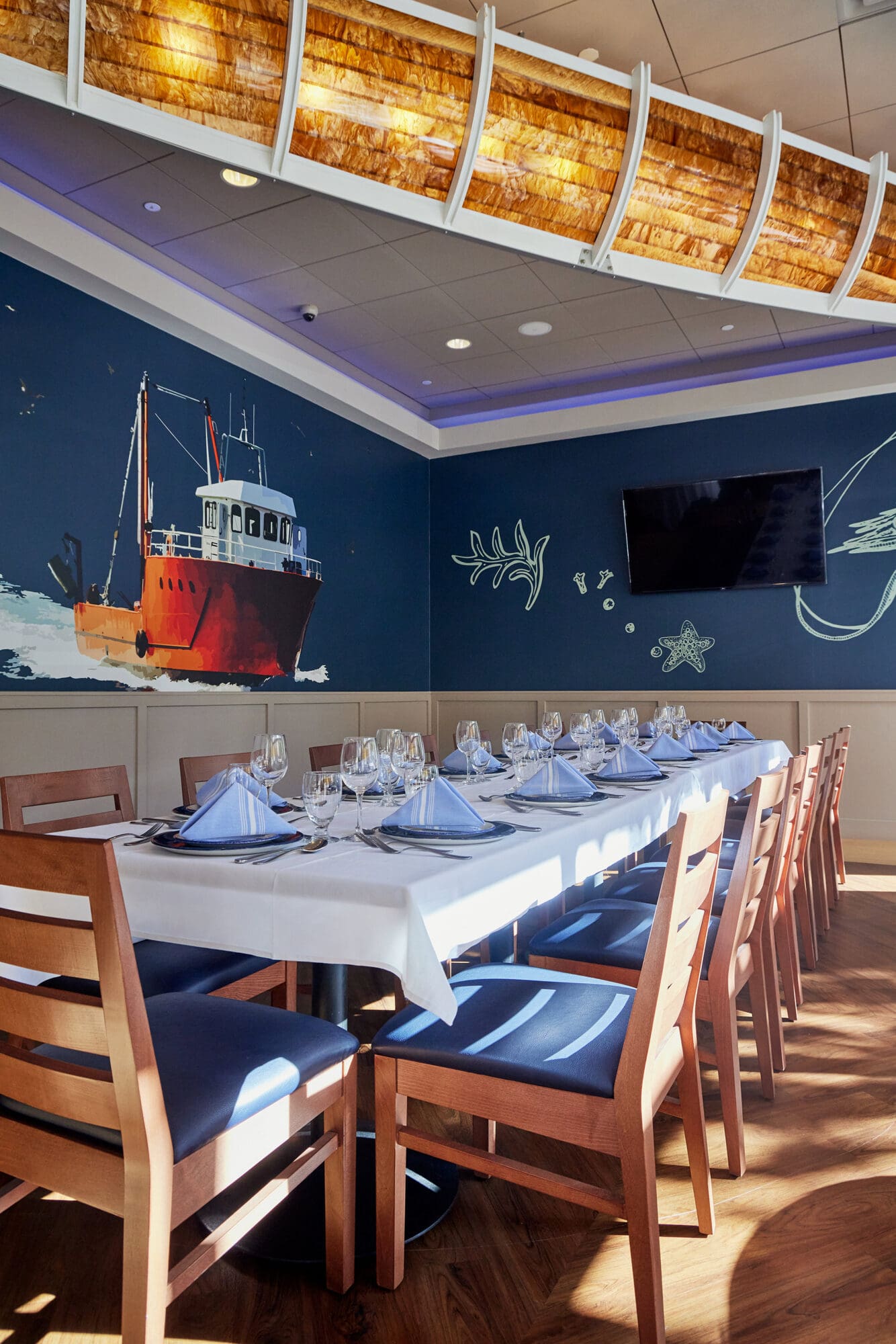 A dining area with a long table set for ten people. Wooden chairs with blue cushions surround the table, which is adorned with neatly folded blue napkins and wine glasses. The wall features a mural of a fishing boat on the ocean, and a flat-screen TV is mounted on it.