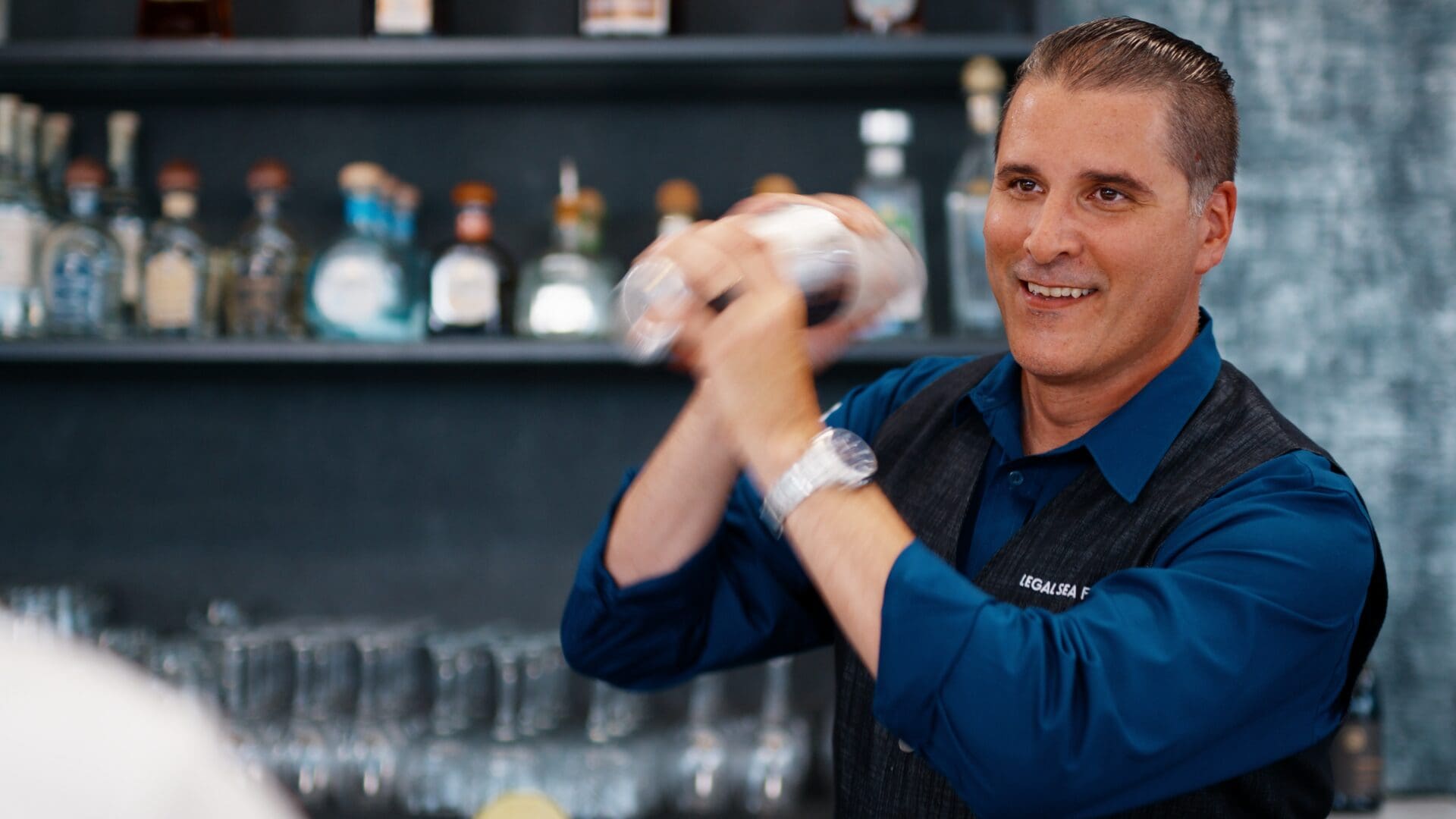 A smiling man is shaking a cocktail shaker behind a bar. He is wearing a blue shirt with rolled-up sleeves and a black vest. The shelves behind him are filled with various bottles and glasses.