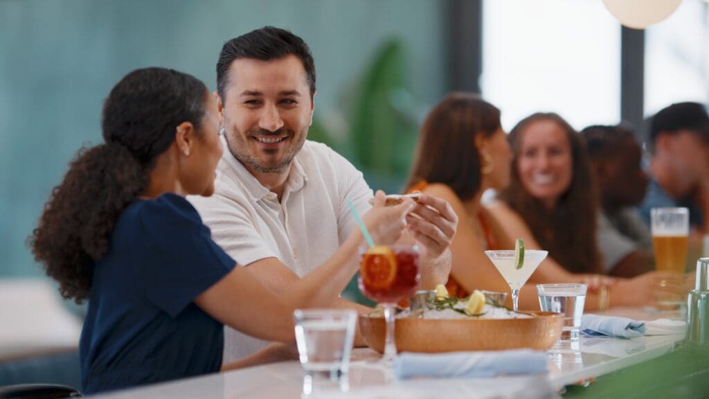 Two people sitting at a bar counter are smiling and talking. One has a cocktail in hand while the other is gesturing mid-conversation. In the background, more people are socializing with drinks and snacks on the counter. The setting is bright and modern.