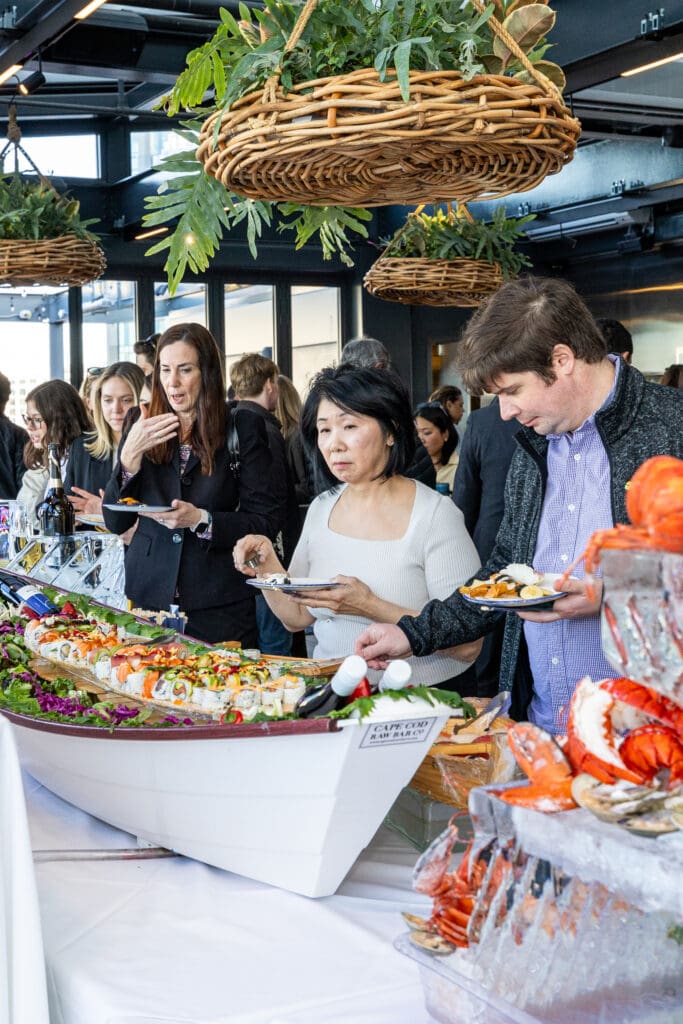 A diverse group of people at a social event is gathered around a long buffet table filled with assorted seafood dishes. Some are serving themselves while others are engaged in conversation. Decorative elements, such as hanging baskets with greenery, adorn the space.