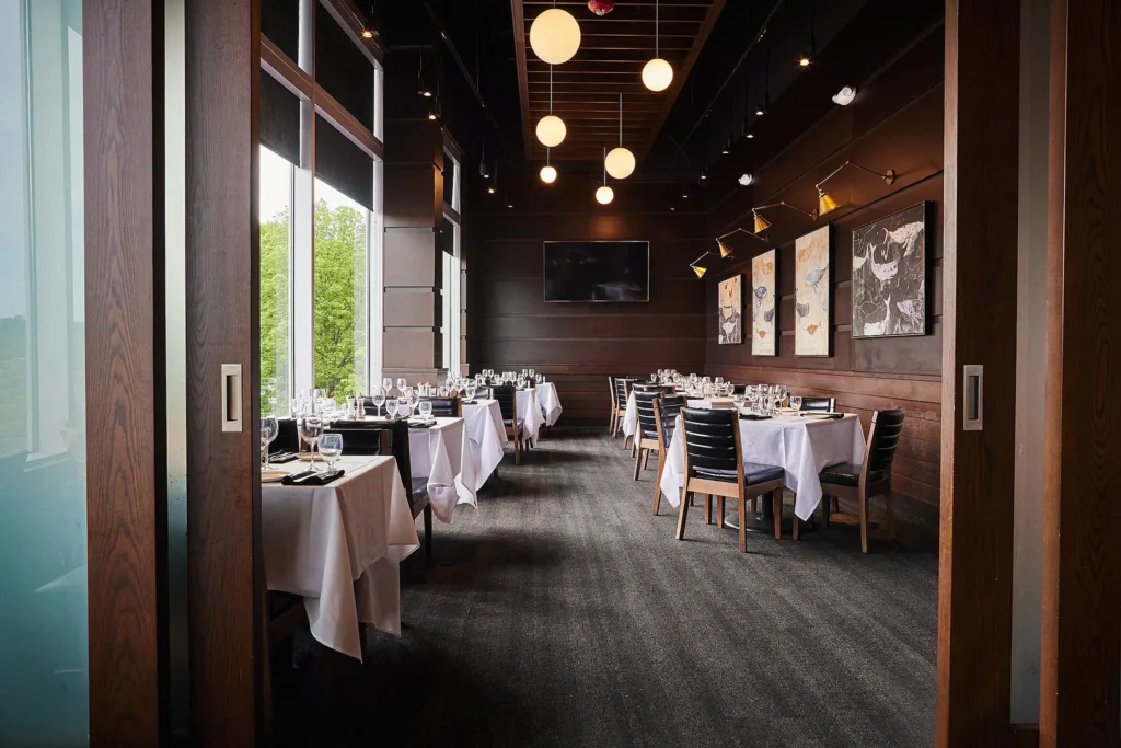 A modern restaurant interior featuring rows of tables set with white linens, glassware, and black chairs. Large windows on the left offer views of greenery outside. Contemporary artwork is displayed on dark wooden walls, and round ceiling lights provide illumination.