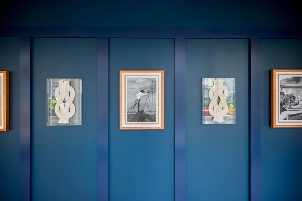 A deep blue wall adorned with three framed artworks. The center frame features a black-and-white photograph of a person on a boat, while the other two frames contain abstract sculptures with vertical, ribbon-like shapes.