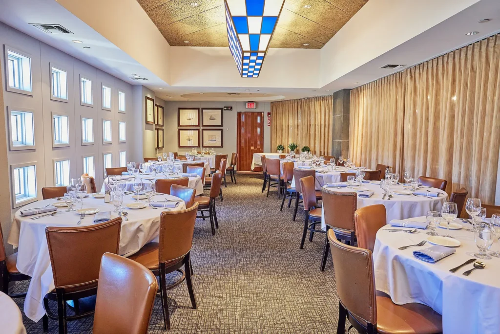An elegant dining room is set with round tables covered in white tablecloths and surrounded by brown leather chairs. Each table is neatly arranged with glassware, silverware, and napkins. Soft lighting and framed artwork on the walls create a warm and inviting atmosphere.