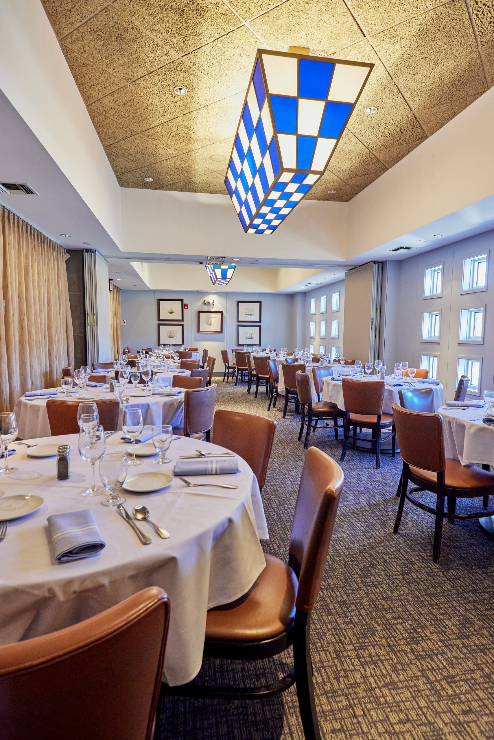 A fine dining restaurant with neatly set tables covered in white tablecloths, brown chairs, and place settings including wine glasses and napkins. The lighting is warm with modern blue and white checkered light fixtures hanging from the ceiling. Framed art adorns the walls.
