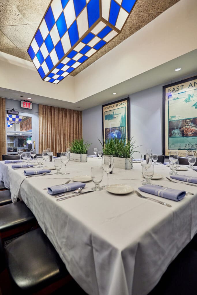 A well-lit dining room features a long table set with white tablecloths, plates, glasses, and napkins. Decorative plants are placed in the center. Blue and white checkered light fixtures hang from the ceiling. Large framed posters adorn the walls.