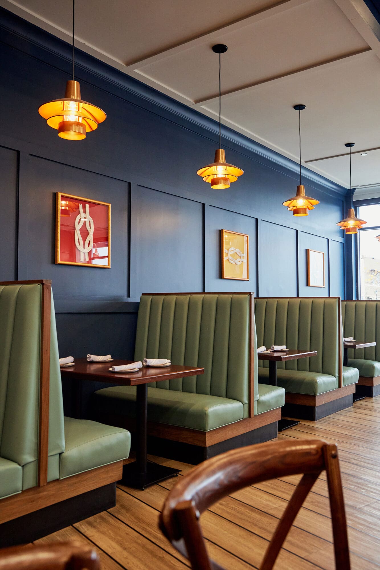 A cozy, modern restaurant interior featuring teal leather booth seating and dark wooden tables. Warm overhead pendant lights hang along the navy blue paneled wall adorned with minimalistic framed art. The floor is lined with wooden planks, and napkins are neatly arranged on the tables.