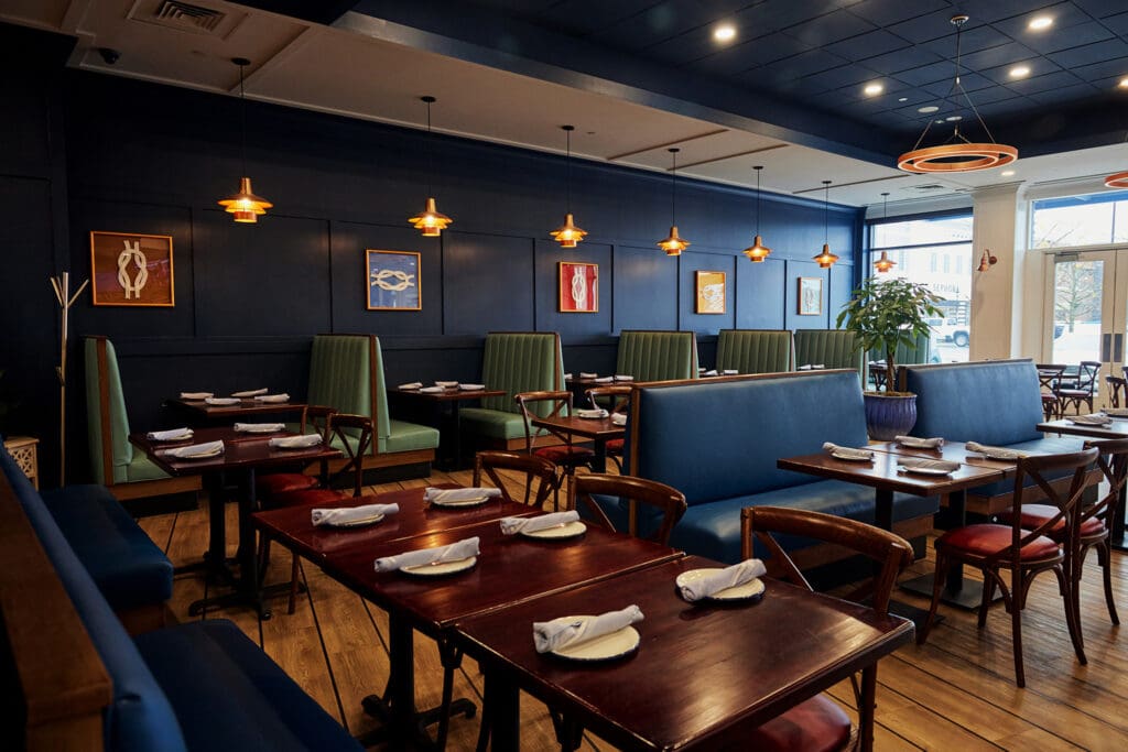 A chic restaurant interior with dark blue walls, wooden floors, and modern light fixtures. Green and blue cushioned booths line the sides, and wooden tables are set with white napkins. Framed art decorates the walls, and a potted plant is near the windows.