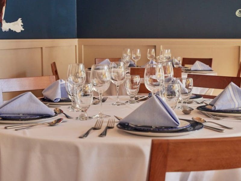 A round table in a restaurant is set for a meal, with neatly folded light blue napkins, clear glasses, and polished cutlery. The table features wooden chairs and is covered with a white tablecloth, against a backdrop of blue and beige wall panels.