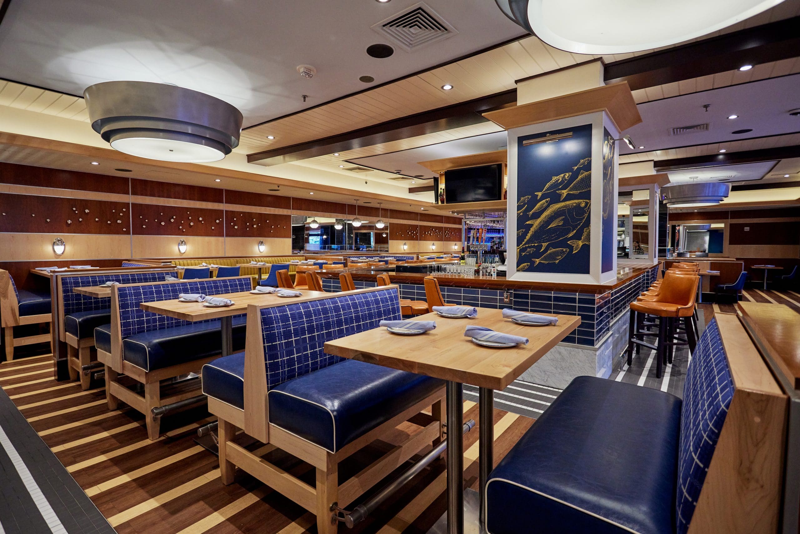 A modern, empty restaurant with wooden decor and navy blue cushioned seating. Tables are set with white plates and napkins. Warm lighting enhances the cozy ambiance. The restaurant features a nautical theme, evident from fish illustrations and maritime accents.