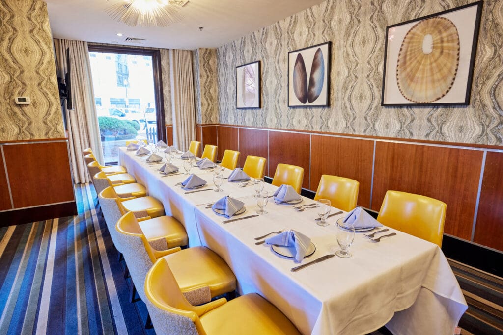 A long dining table with a white tablecloth is set with folded gray napkins, glassware, and silverware in an elegant room. Yellow chairs line both sides of the table. The walls have patterned wallpaper and are adorned with framed art. A high window offers natural light.