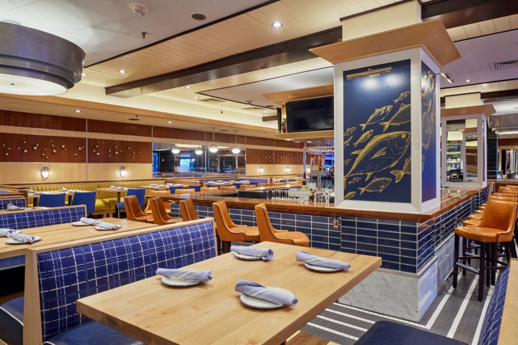 A modern restaurant interior featuring wooden tables with blue-checkered booth seating, neatly arranged napkins, and high chairs along a bar counter. The decor includes a column adorned with fish illustrations and a combination of wood and blue-tiled finishes.